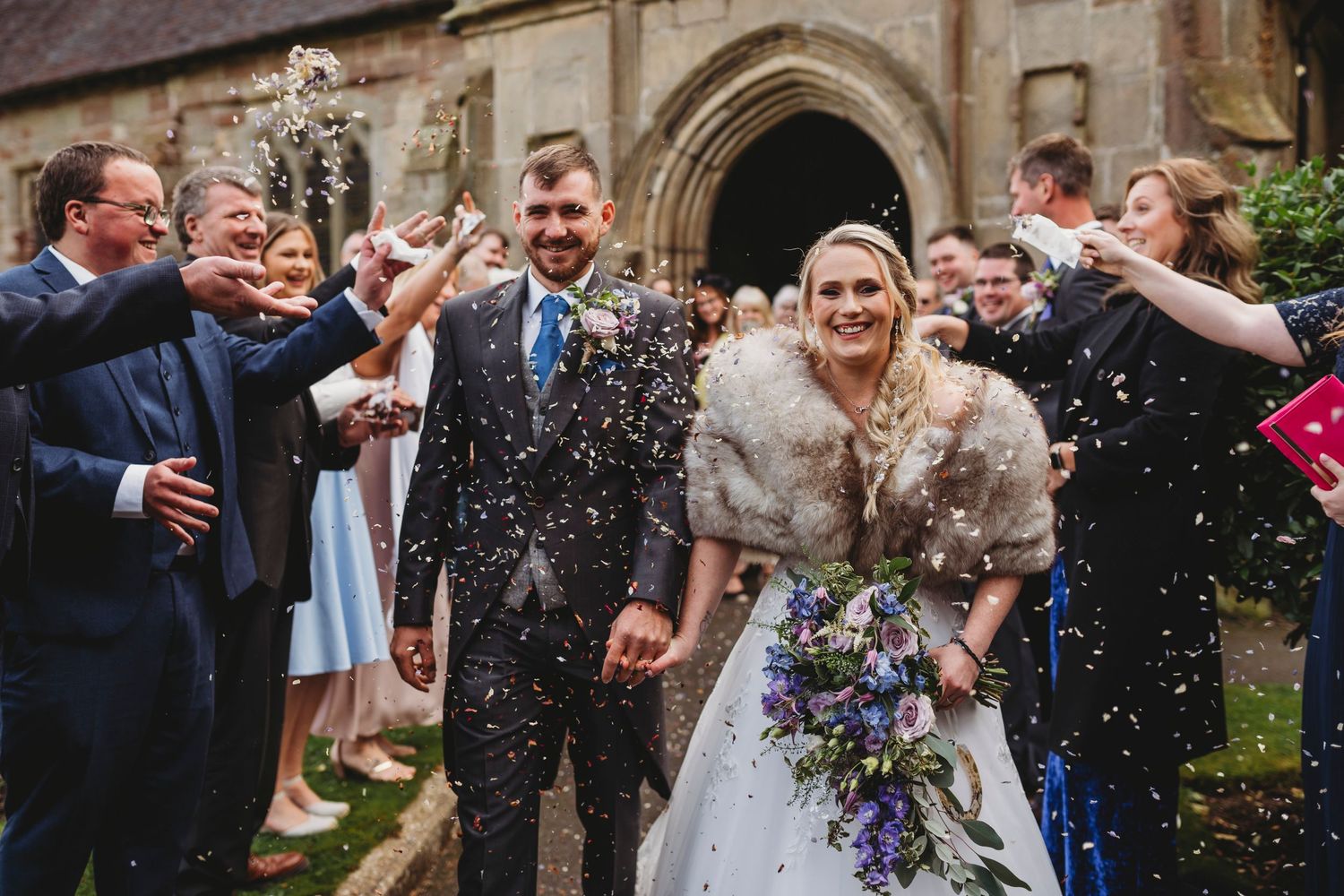 Wedding celebration outside historic stone church with guests throwing confetti.