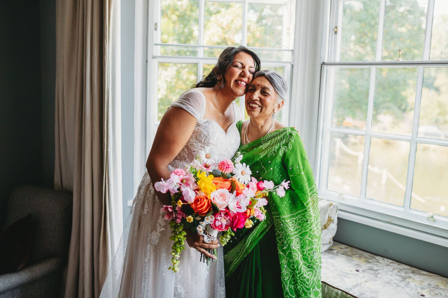 Bride in white gown holds colorful bouquet beside woman in green sari, smiling near bright windows at Morden Park House London