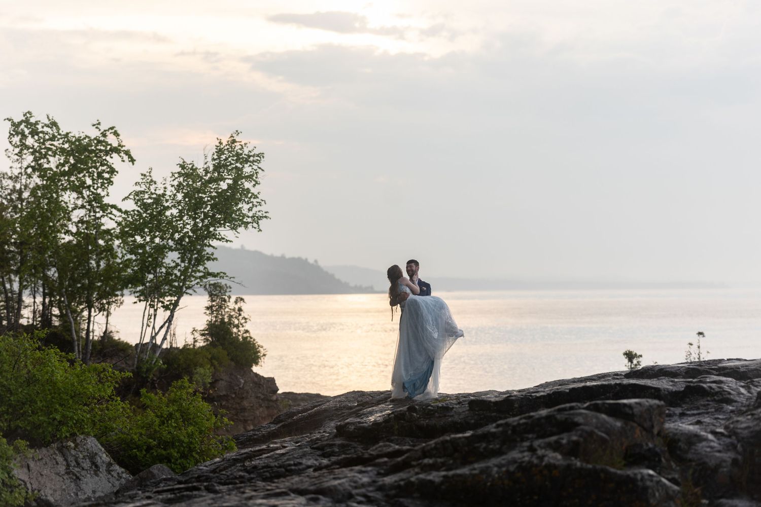 A couple stands on rocky cliffs holding each other at sunset over a serene lake.