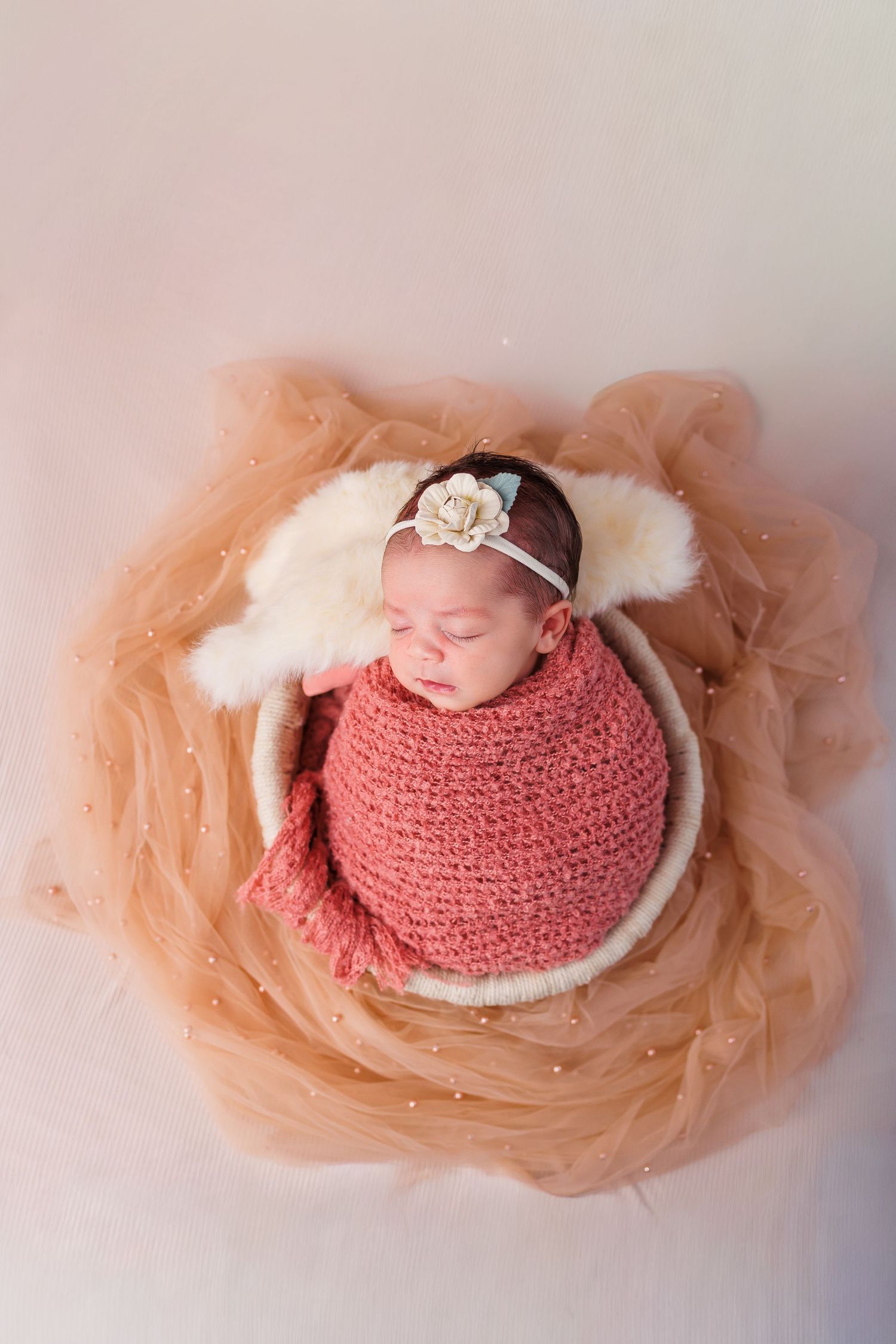 Sleeping newborn baby wrapped in white lace swaddle with small bow headband in cream basket.