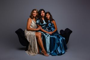Three women in elegant evening gowns pose together on a black velvet sofa against a dark gray background.