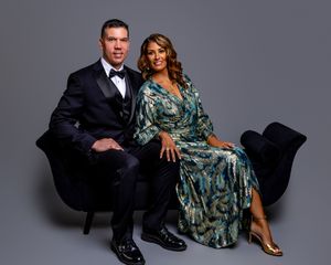 Studio portrait of couple on black velvet sofa, wearing formal attire with navy suit and green floral gown.