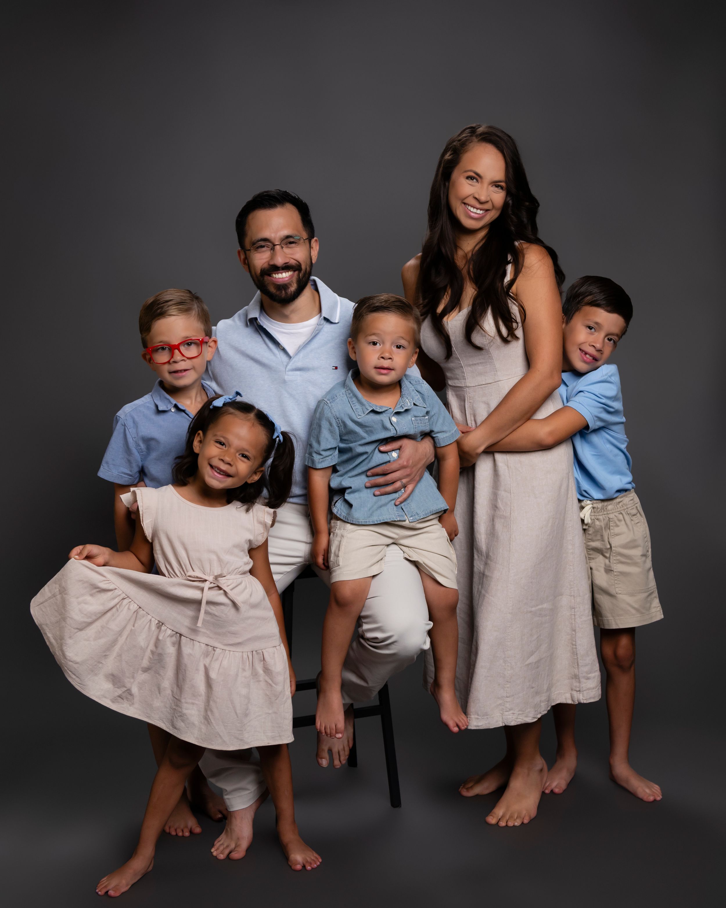 A family of six wearing coordinated neutral outfits poses together against a dark backdrop in a studio portrait.