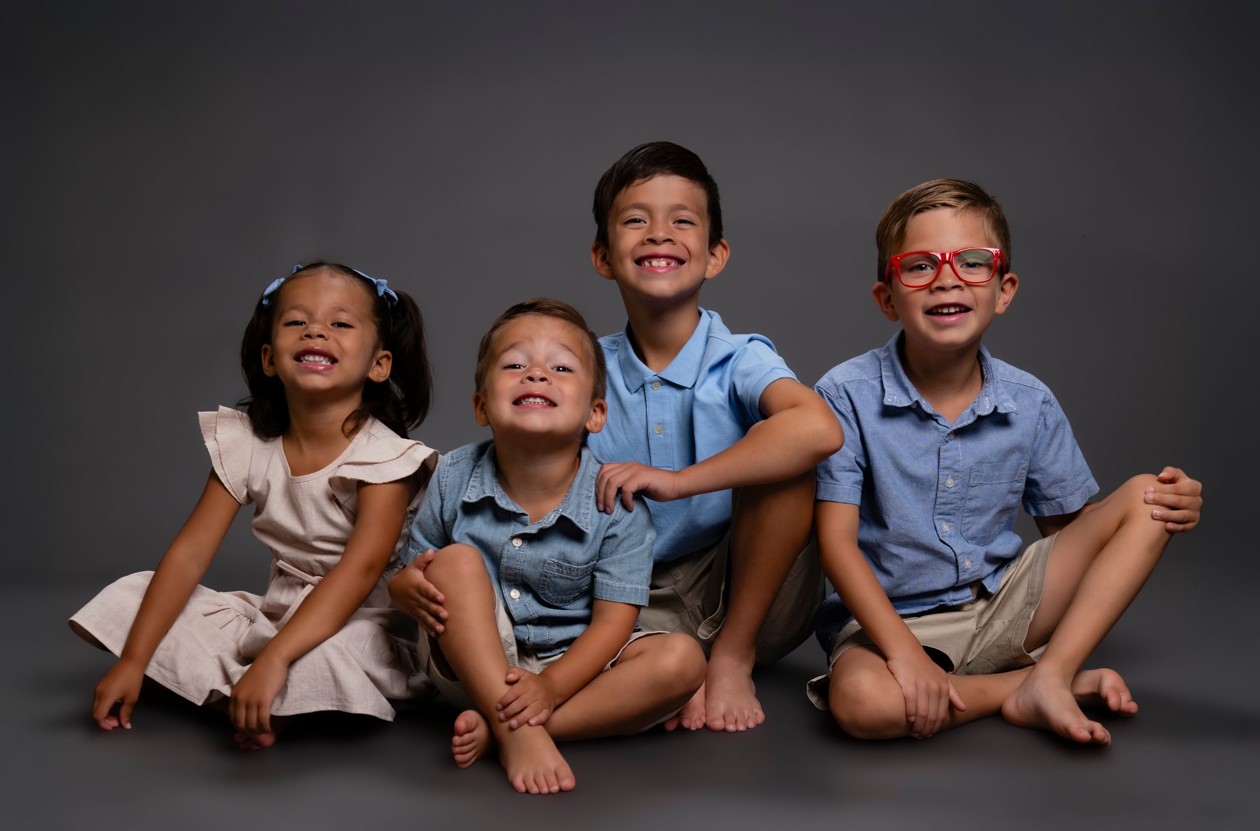 Five siblings sit in a row on a gray backdrop while showing expressions of joy and laughter.