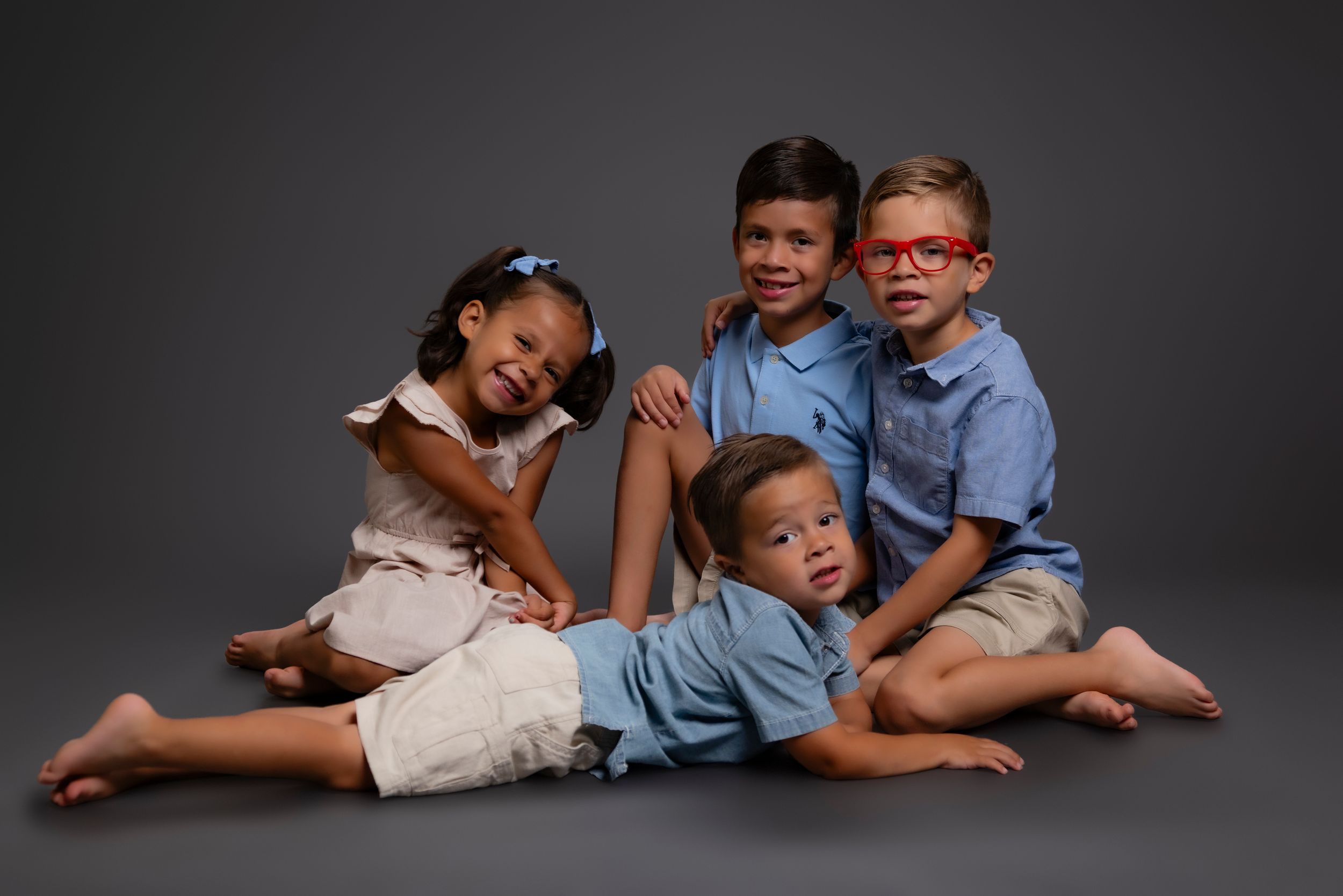 Four young children pose together on a dark gray backdrop wearing coordinated outfits in blue and beige tones.