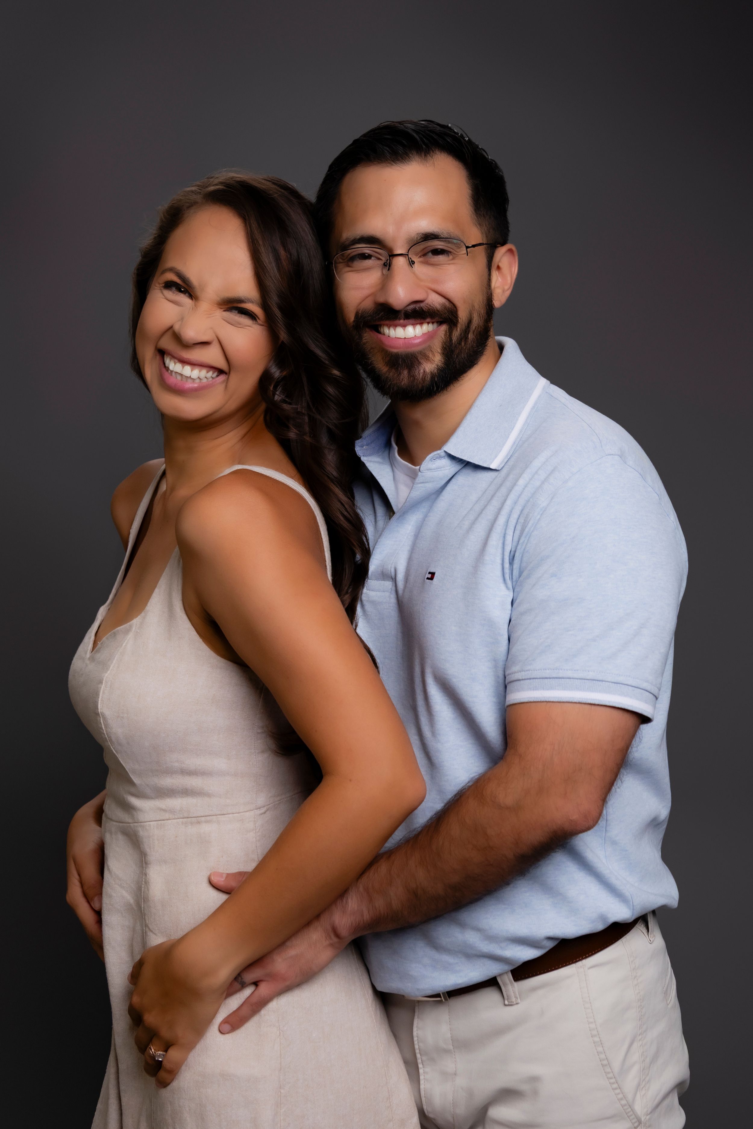 A couple stands back-to-back while smiling warmly against a dark background in a professional studio portrait.