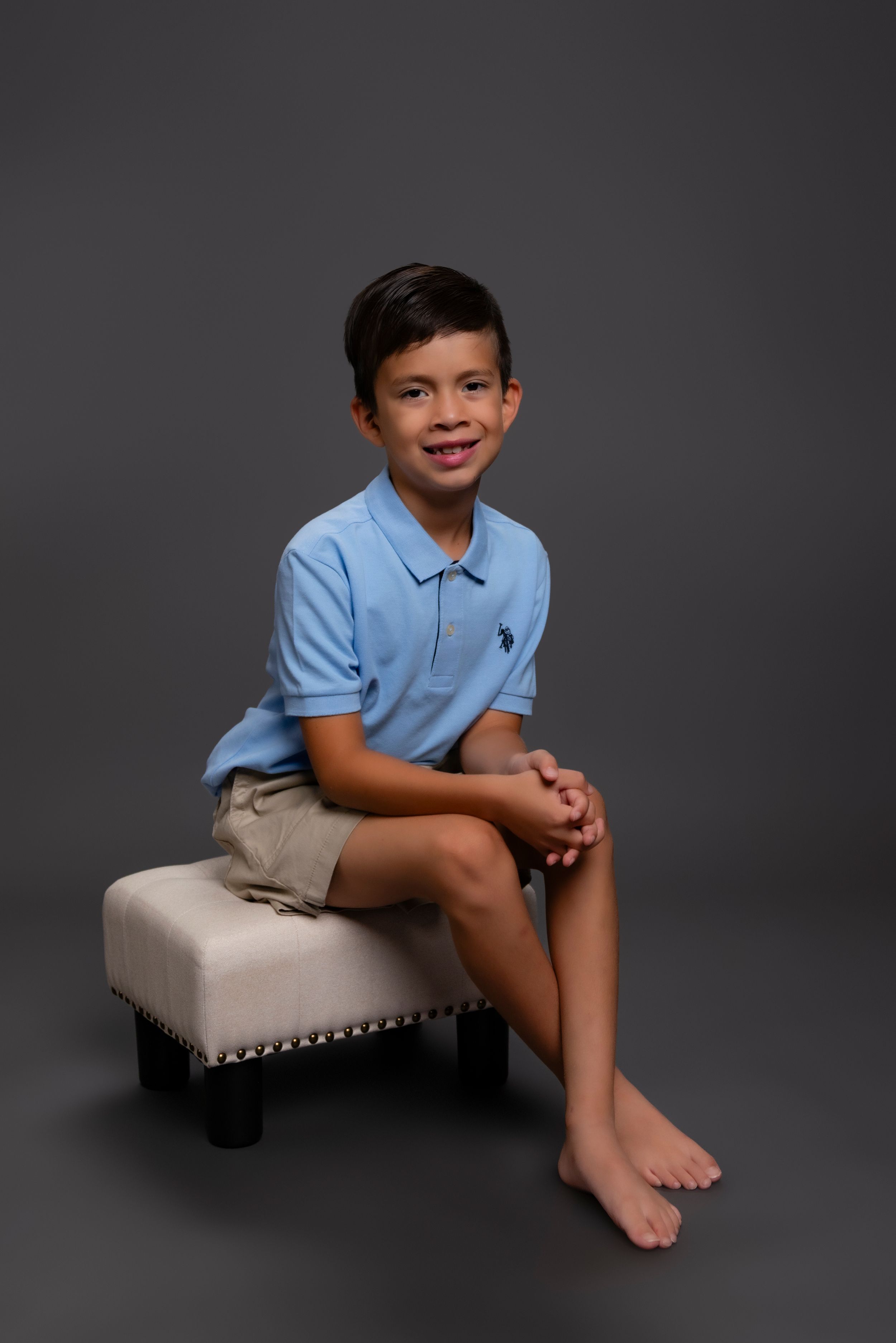 A young boy sits barefoot on a cream-colored ottoman wearing a light blue polo shirt and khaki shorts in a studio setting.