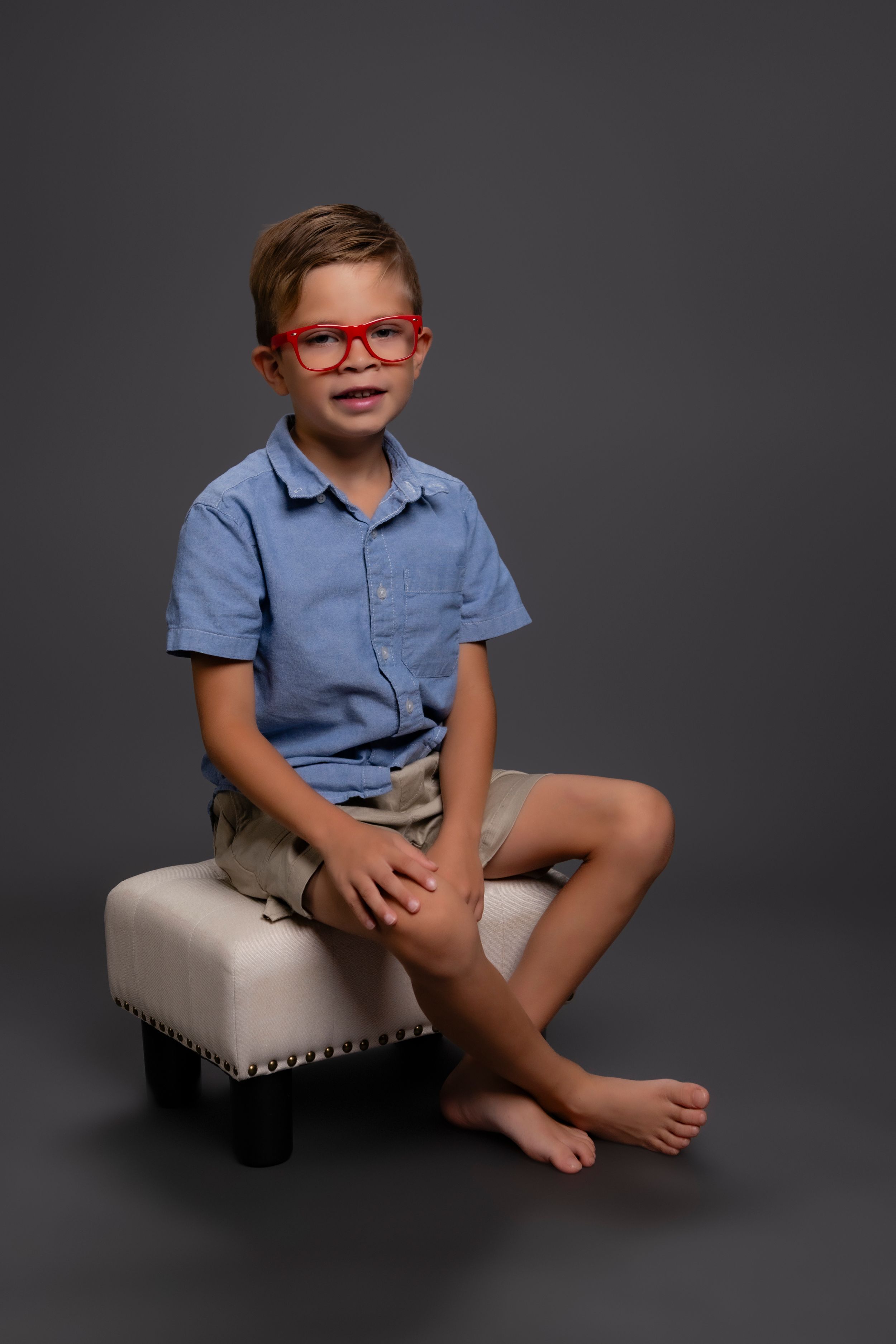 A child in bright red glasses and a blue shirt sits barefoot on a light-colored upholstered ottoman against a dark backdrop.