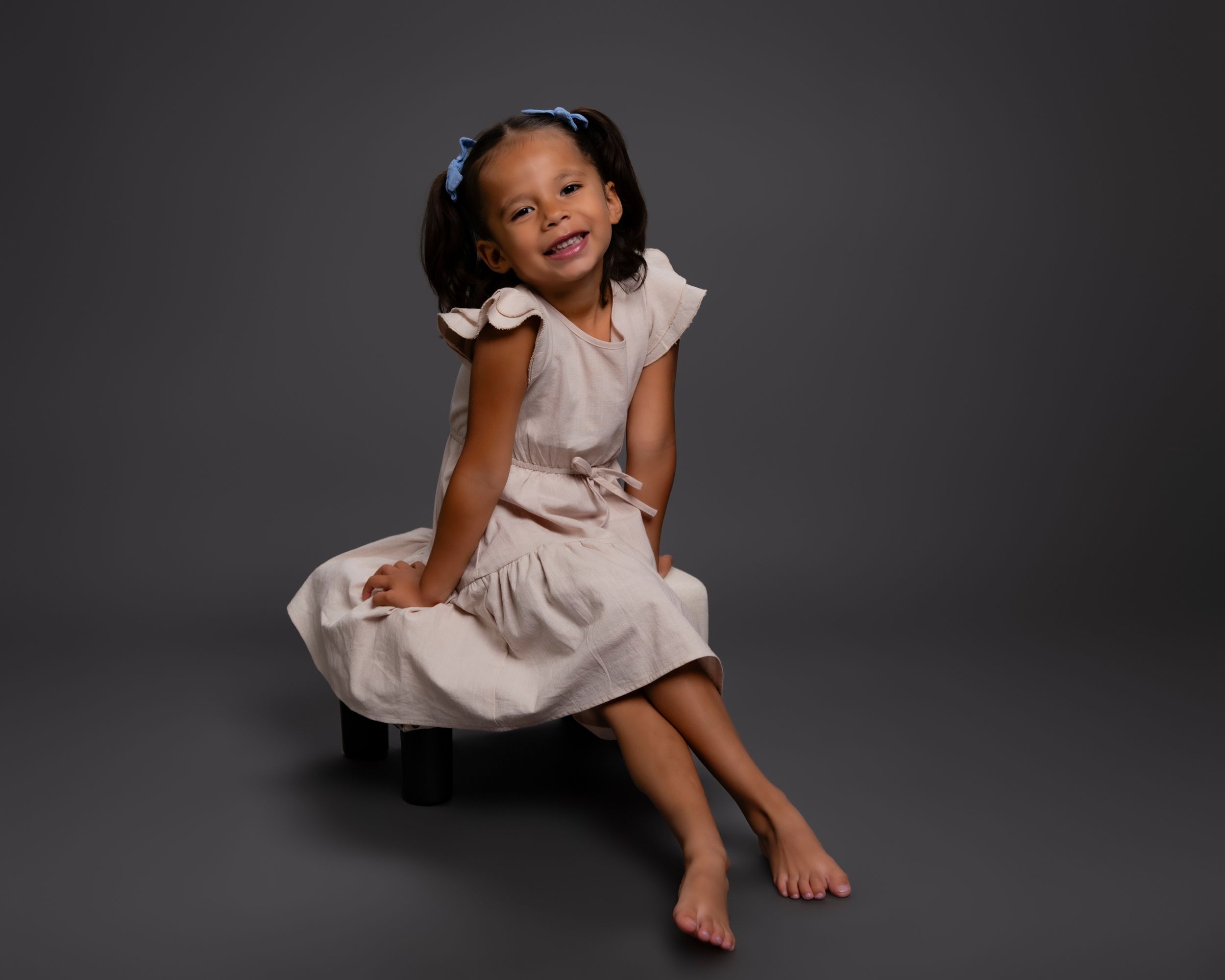 Young child in beige dress sits barefoot while smiling against dark background during professional studio photoshoot.
