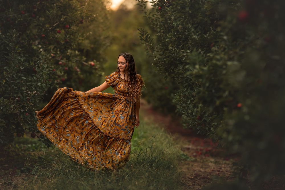 Woman in a golden dress twirls and dances through an orchard path at sunset.