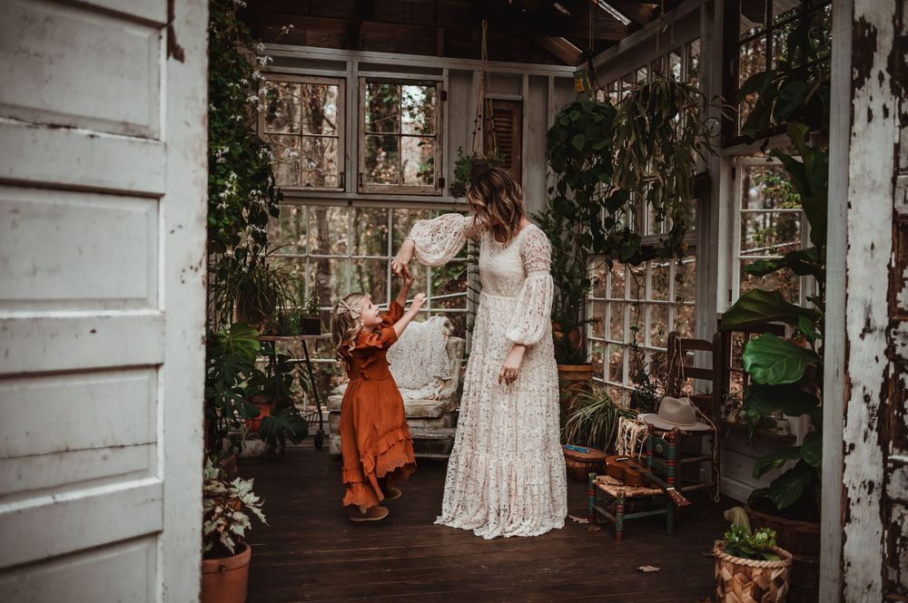 A vintage marietta greenhouse decorated with plants and antiques features a mother in a white lace dress and daughter in an orange dress.