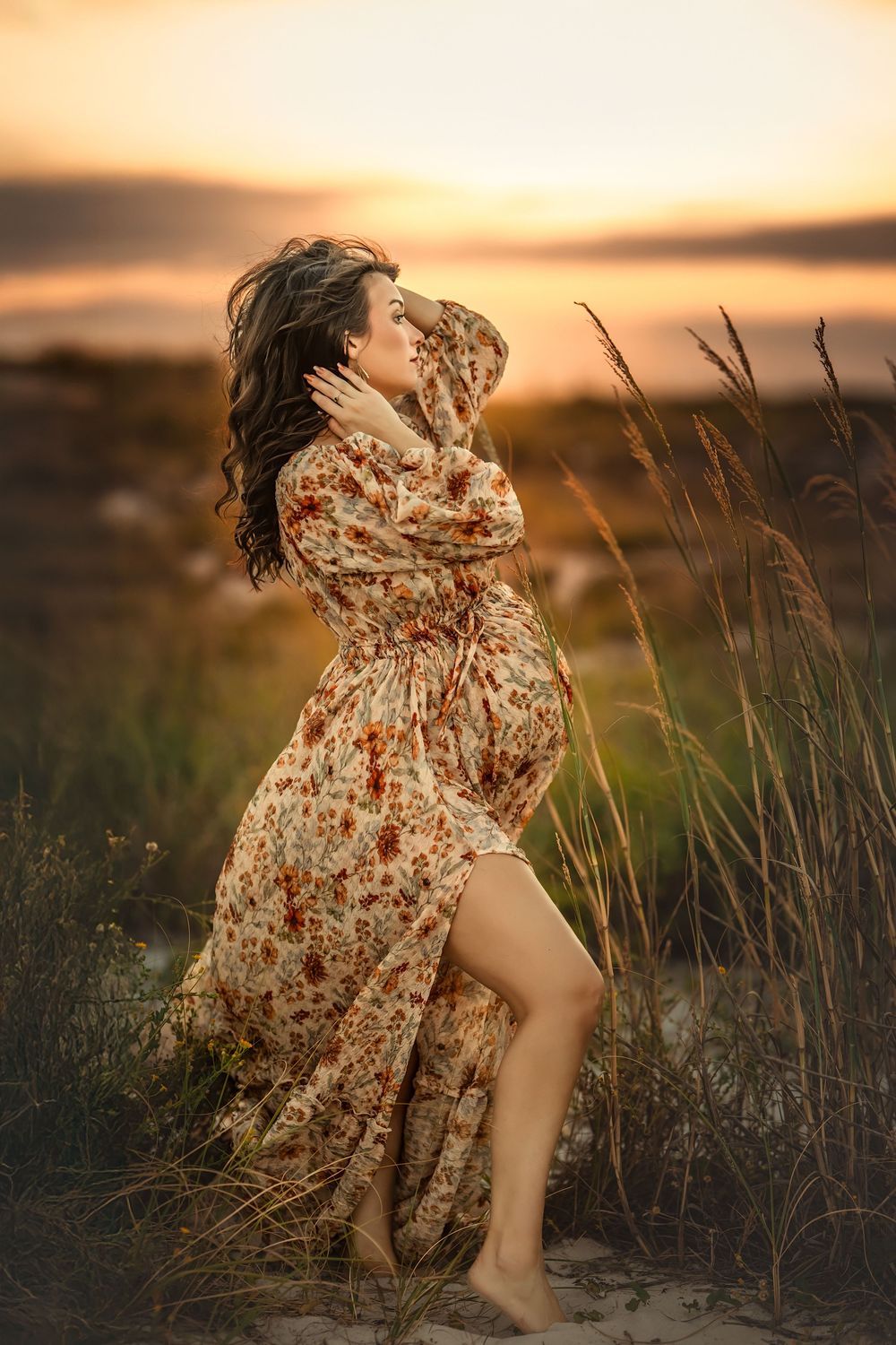 A pregnant woman in a floral dress dances through tall grass during golden hour sunset.