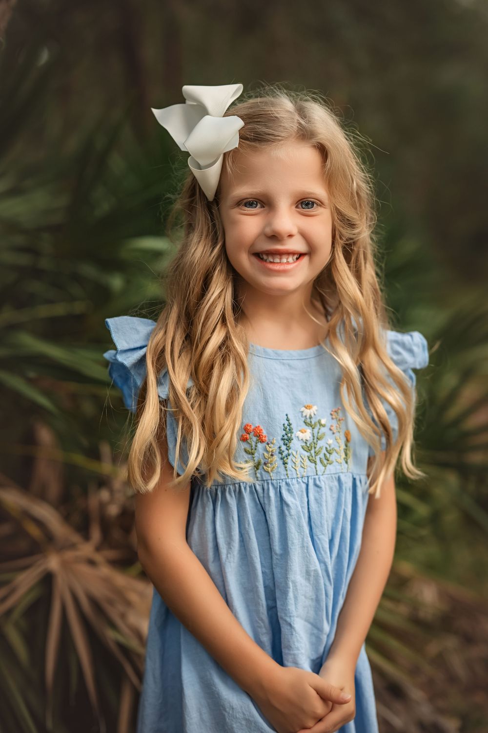 A child in a light blue dress with embroidered flowers stands in front of lush greenery.