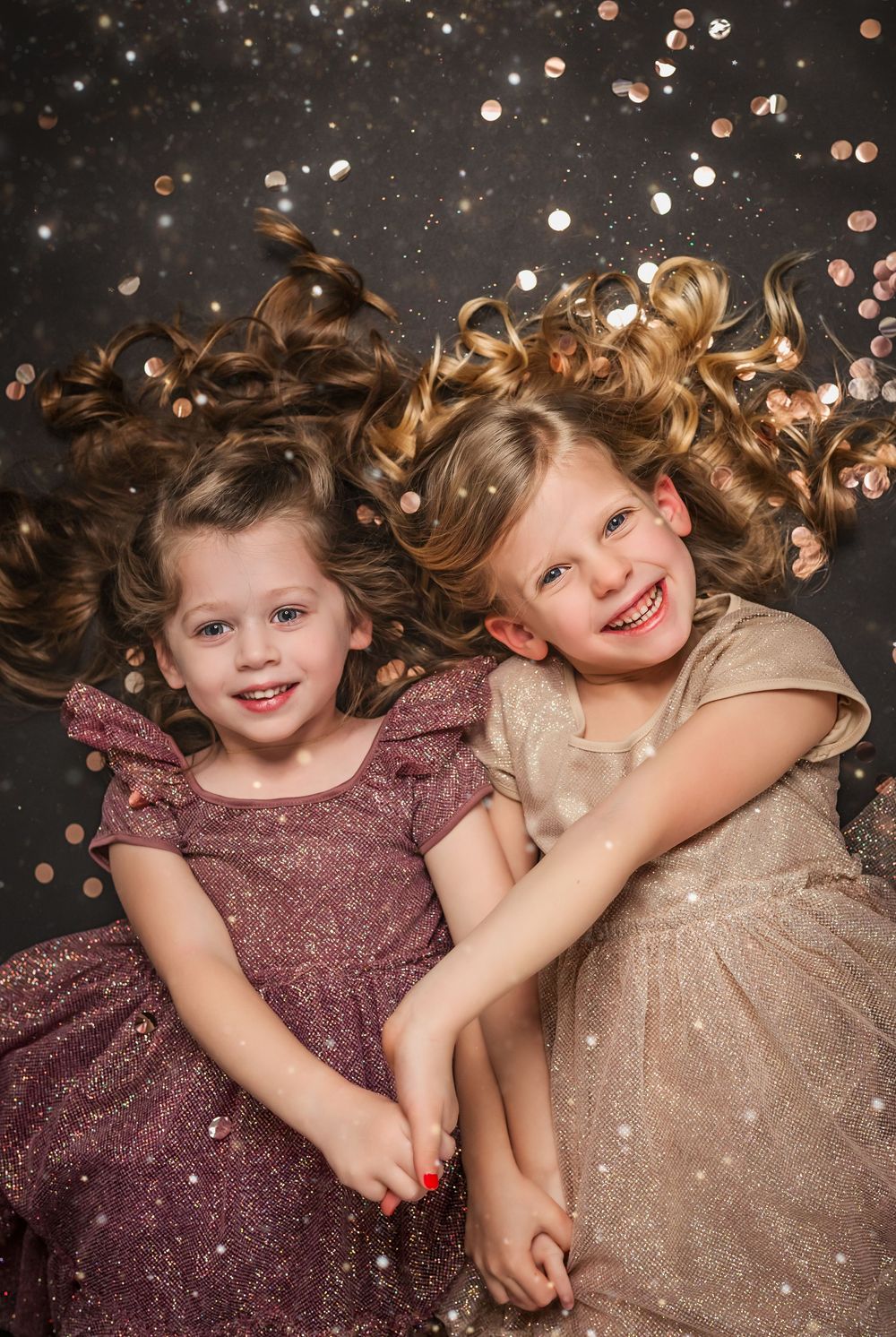 Two young children in sparkly dresses lying down with curly hair spread out among glittering confetti on dark background.