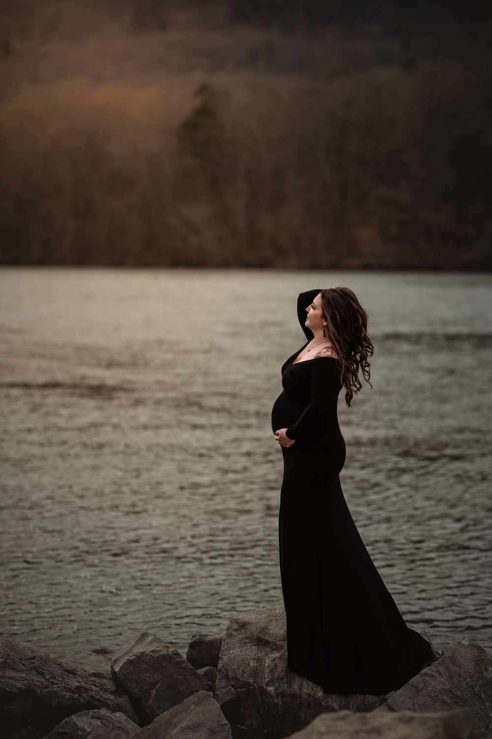 A silhouetted pregnant woman in black stands on rocky shoreline at dusk against dark moody waters.