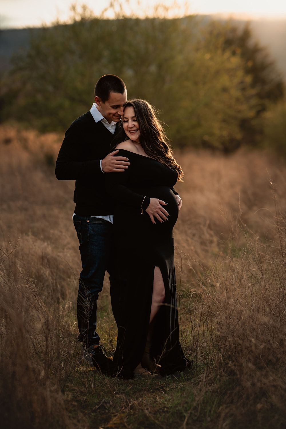 A couple embraces in a field during sunset, wearing black and dark clothing.