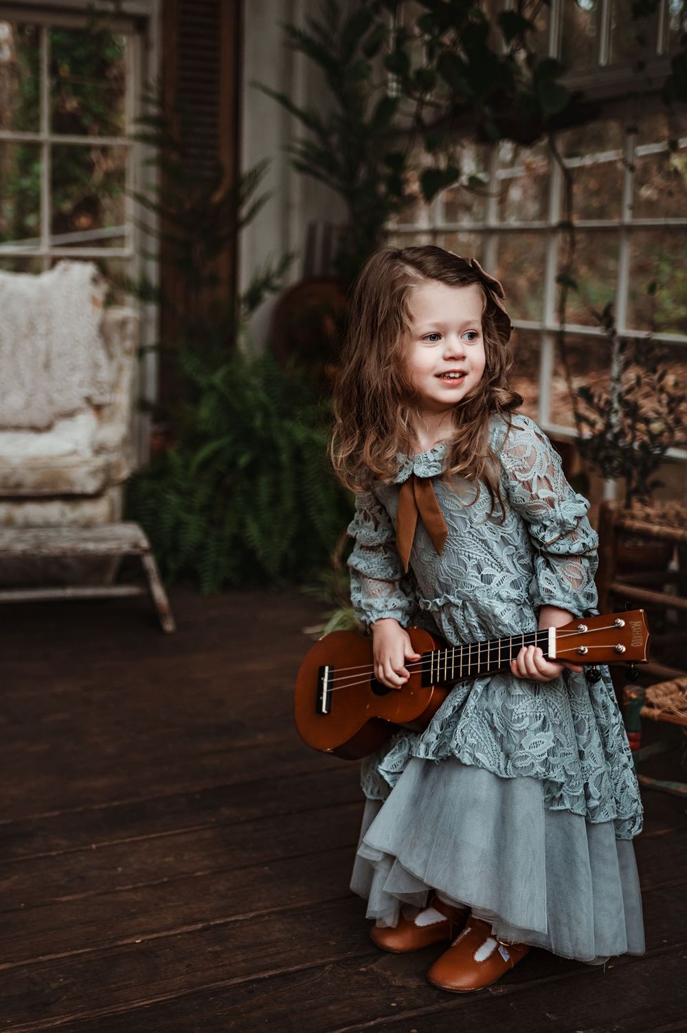 A small child in a ruffled blue dress plays a ukulele while sanding on a dark wooden floor.