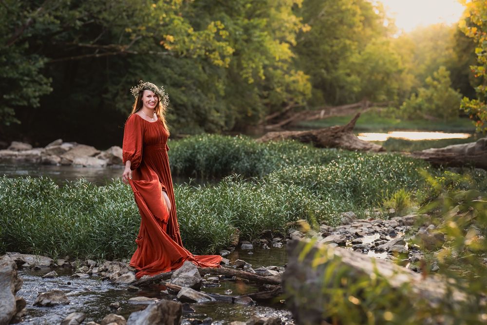 A woman in a flowing red dress stands gracefully in a stream surrounded by lush greenery at golden hour.