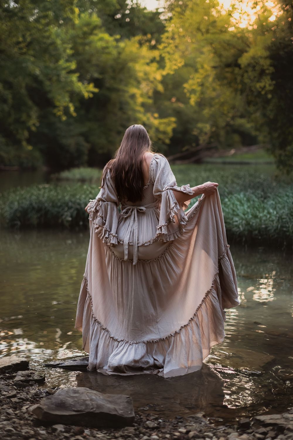 A woman in a flowing vintage dress stands on rocks by a creek surrounded by summer foliage.