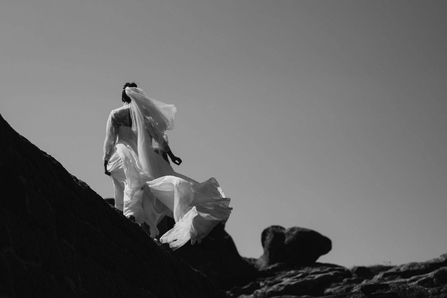 Black and white dramatic shot of bride in flowing gown standing on rocky landscape against clear sky.