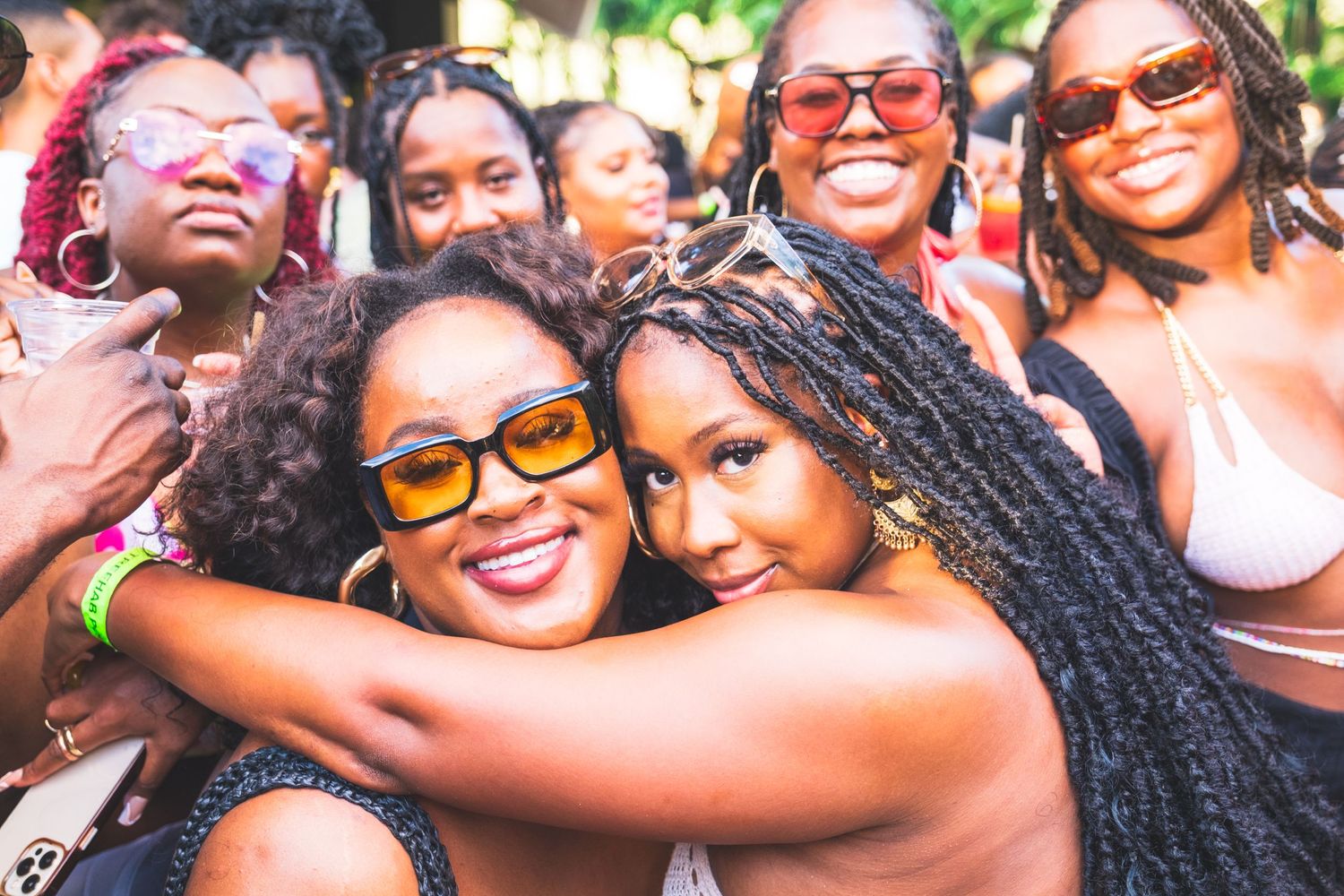 A group of friends wearing sunglasses smile together while taking a selfie at an outdoor gathering.