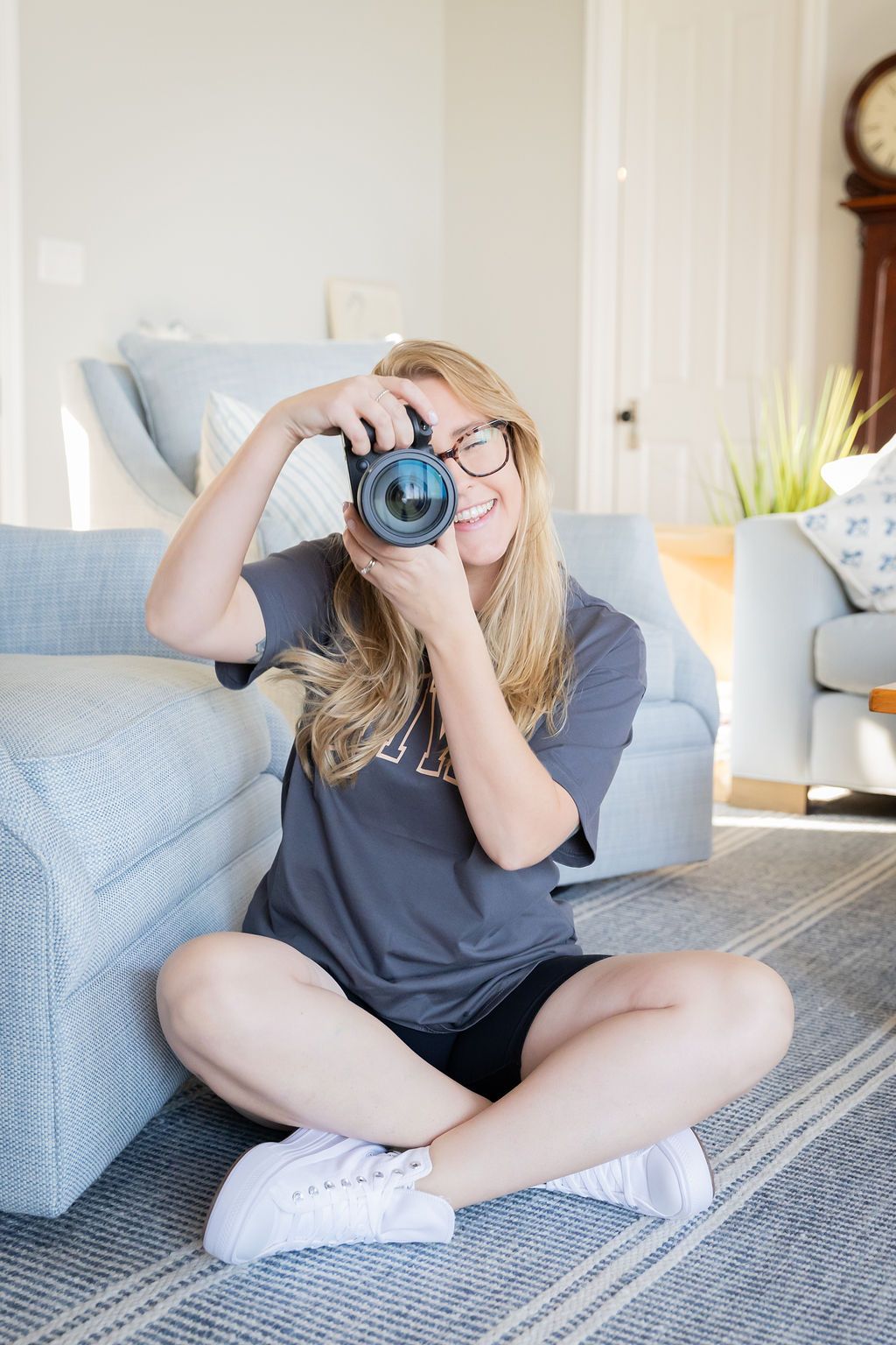 A photographer holds up a camera while sitting cross-legged on a grey area rug.