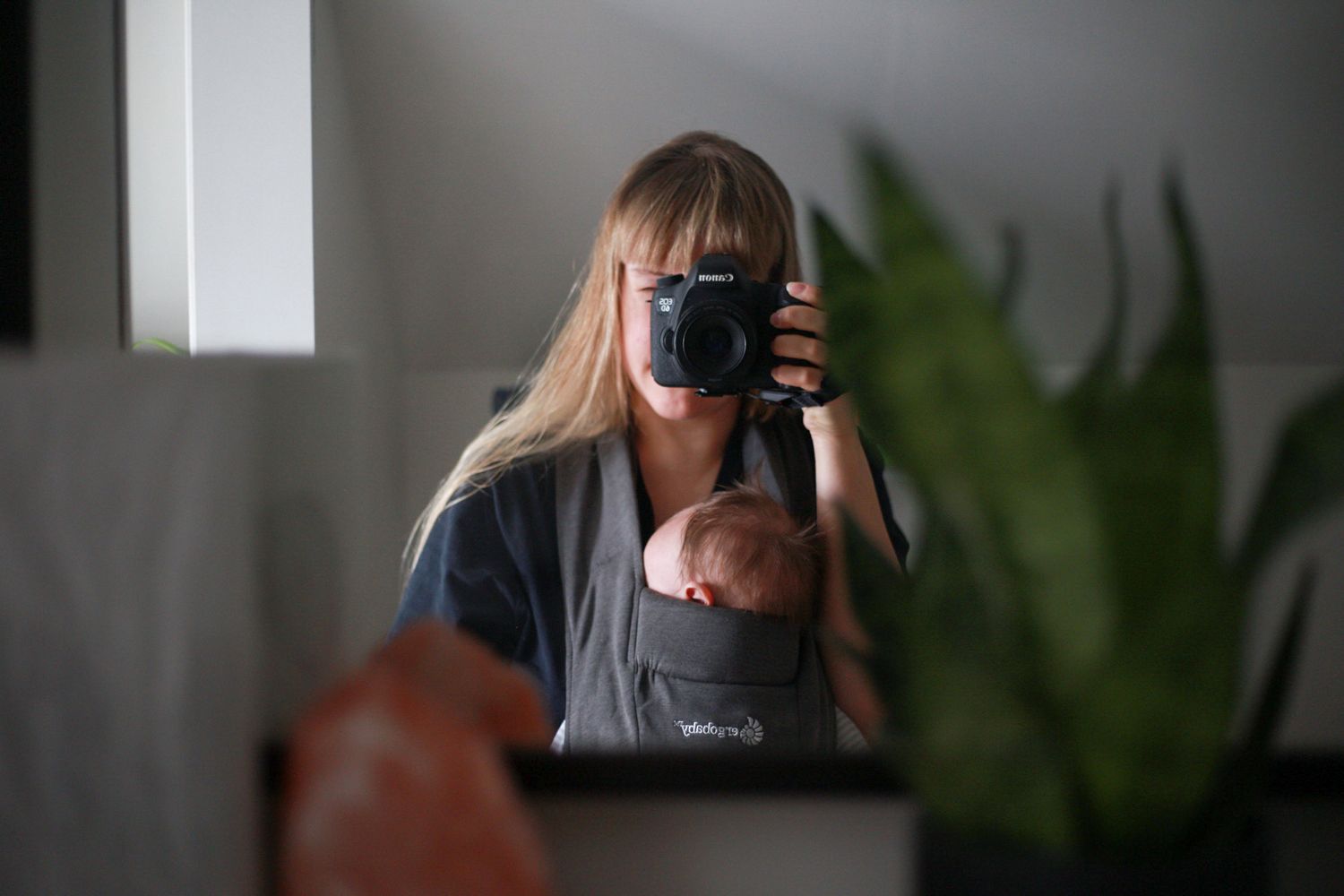 A photographer takes a self-portrait in a mirror showing a camera and plant in the background.