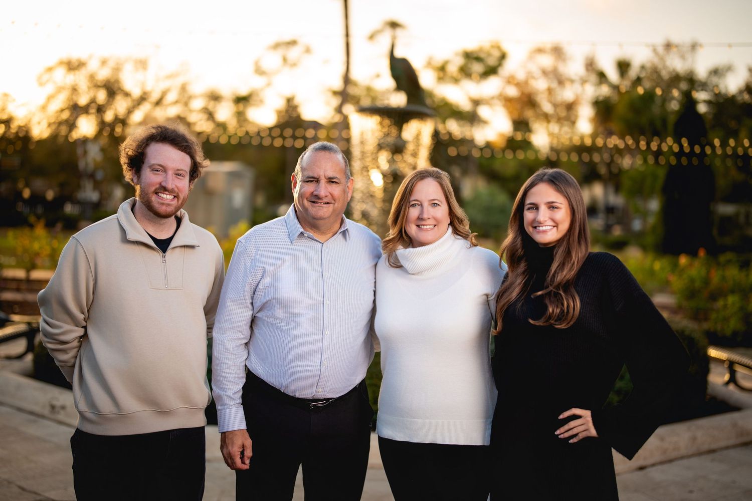 Four people stand together outdoors at sunset wearing casual attire with warm lighting in the background.