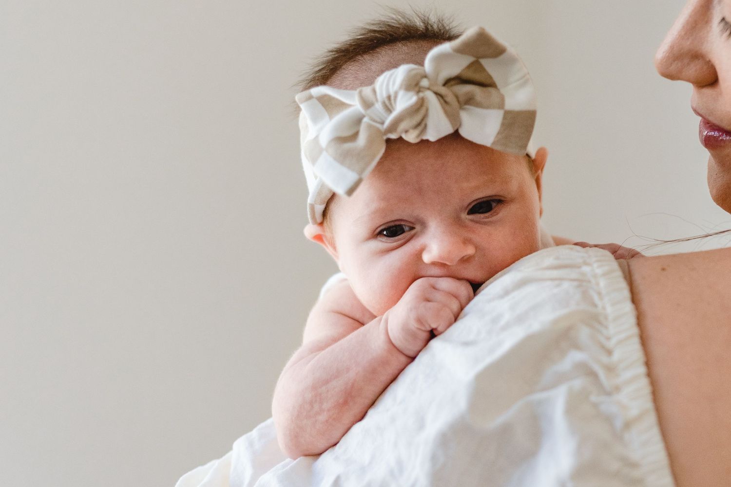 Baby wearing a bow headband rests against a shoulder in soft natural light.