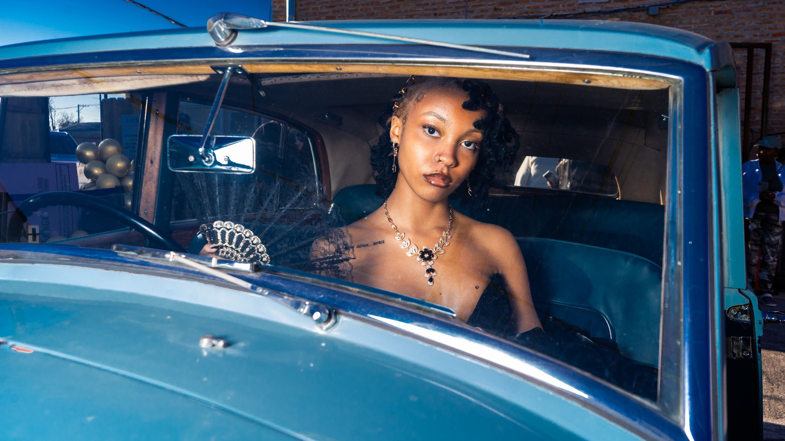 Person with tattoos and jewelry sitting in a vintage blue car on a sunny day.