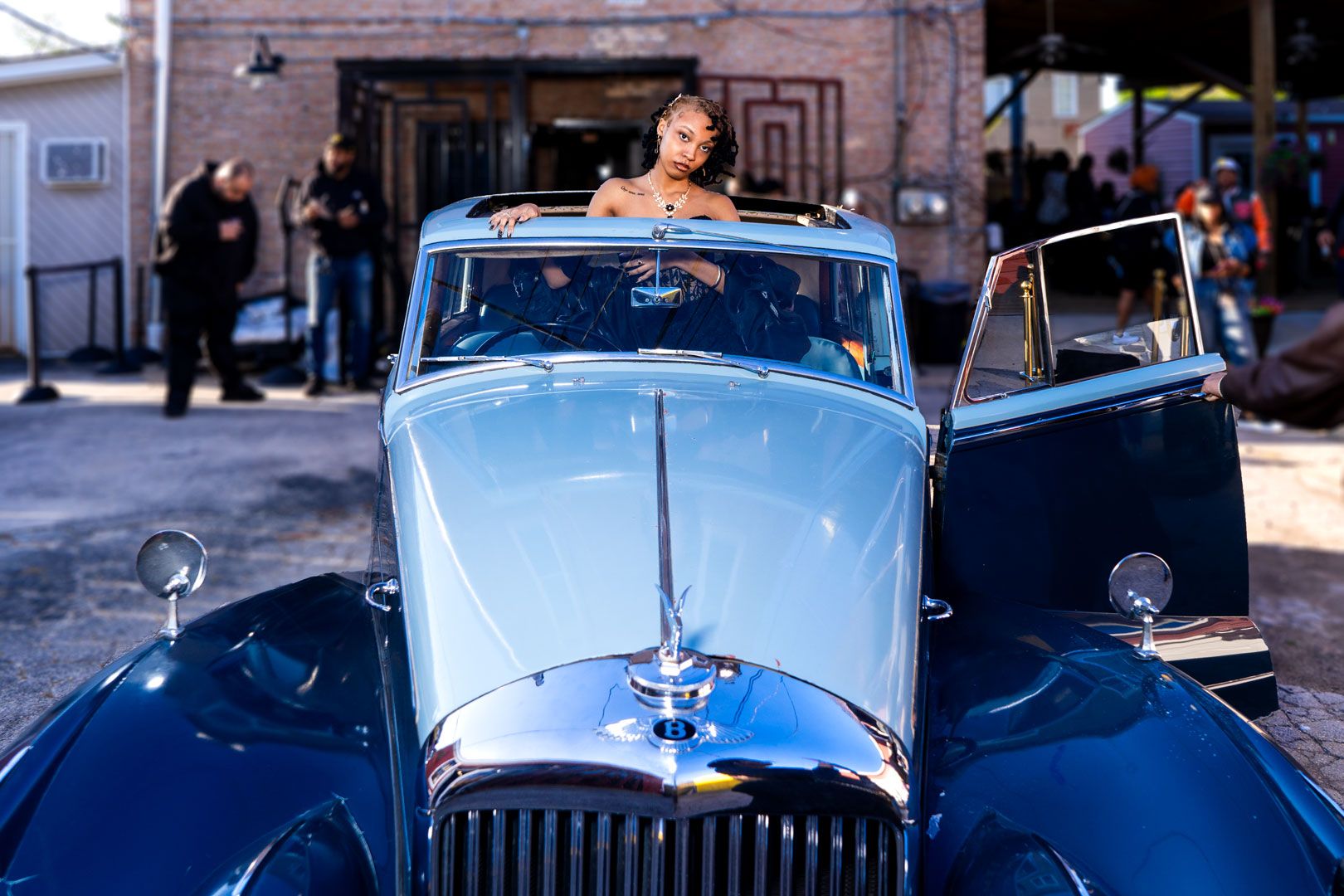 A vintage blue luxury car with chrome hood ornament and grill parked on a sunny street.