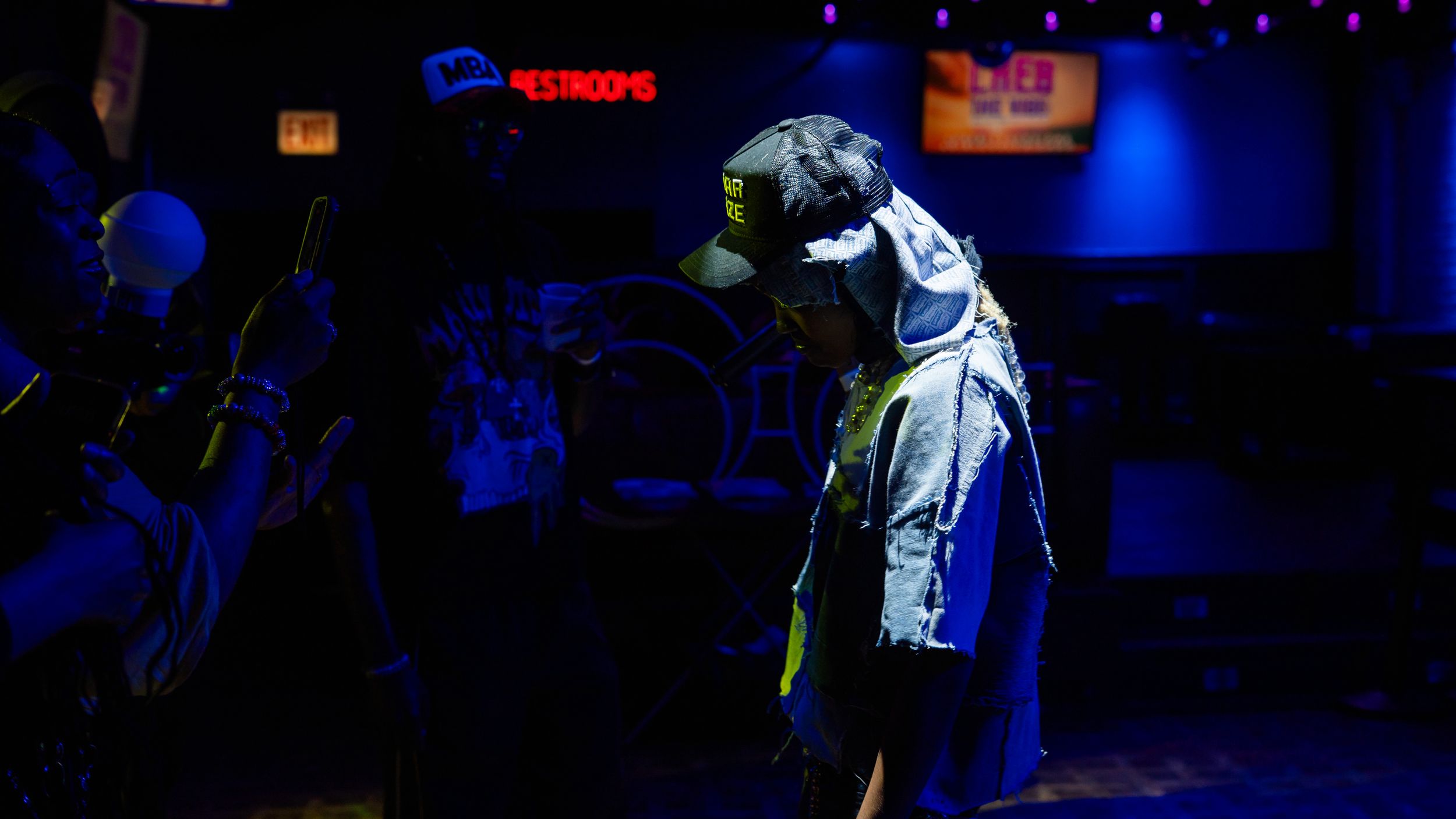 A performer in a white jacket stands illuminated on a dark stage with blue lighting in the background.