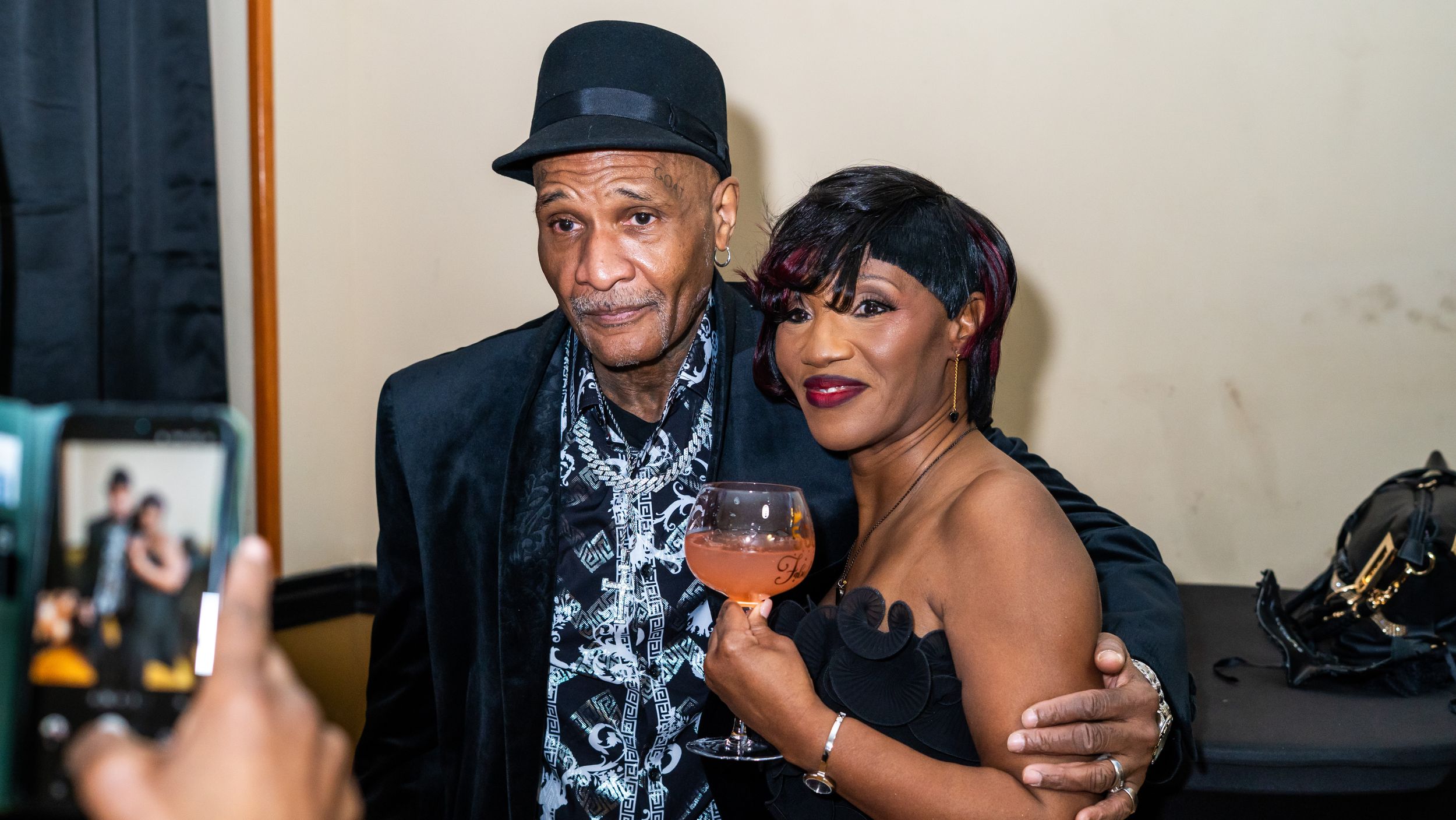 Two elegantly dressed people posing together at a formal event, man in hat and woman in black dress holding a drink.