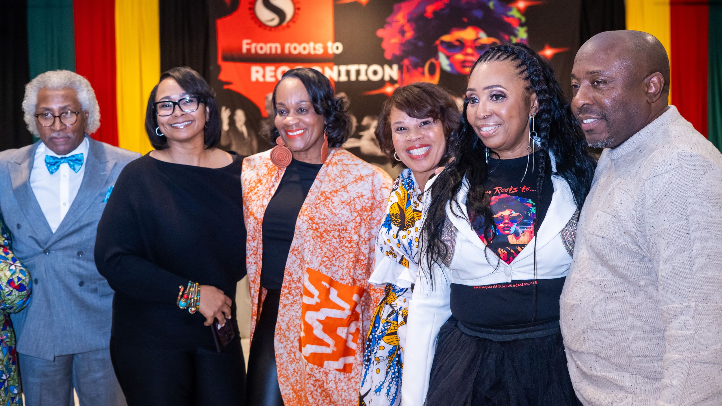 Six people posing together at a From Roots to Recognition event, dressed in colorful formal attire.