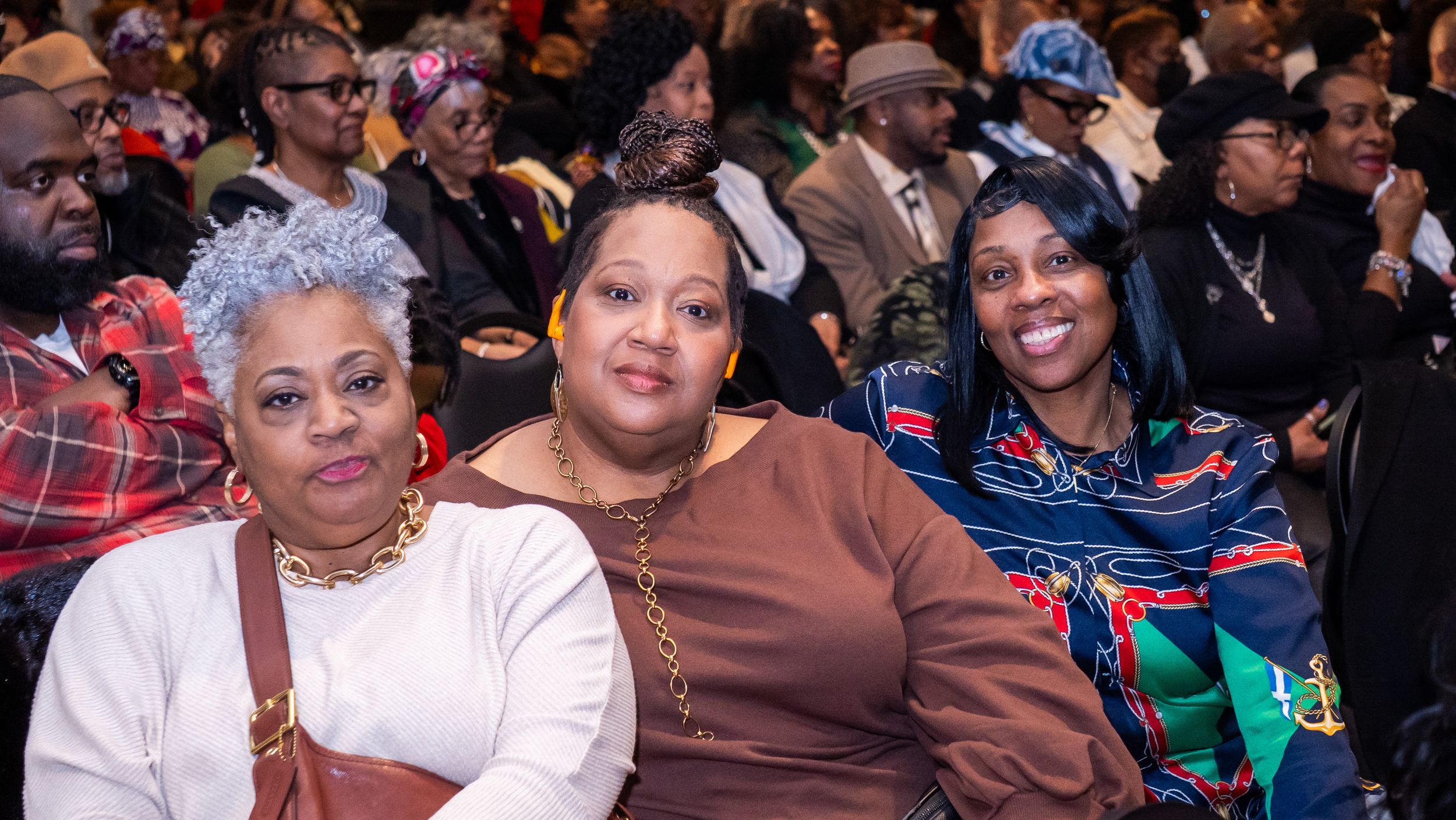 Three women smiling and posing together at a formal indoor event, seated in an auditorium filled with attendees.