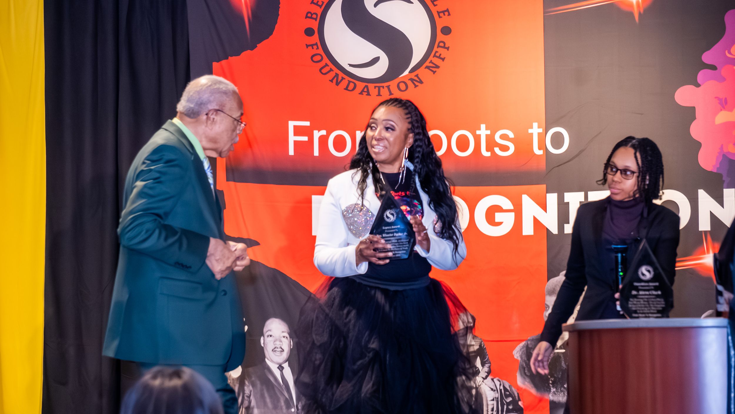 Three people on stage at a Foundation event with a 'From Roots to Recognition' banner backdrop.