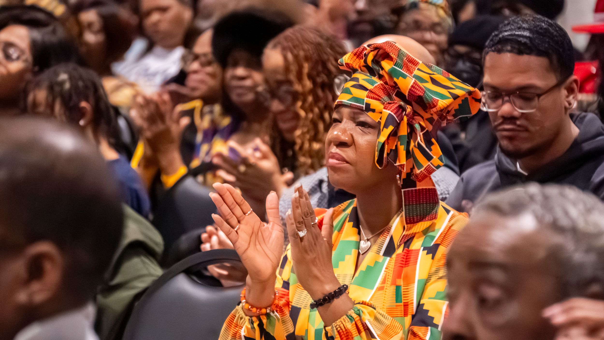 Woman in colorful kente cloth head wrap and dress clapping at a cultural celebration event with diverse crowd.