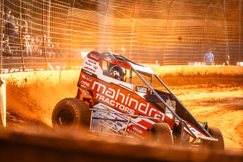 Off-road racing vehicle throwing up dirt on illuminated orange track at night.