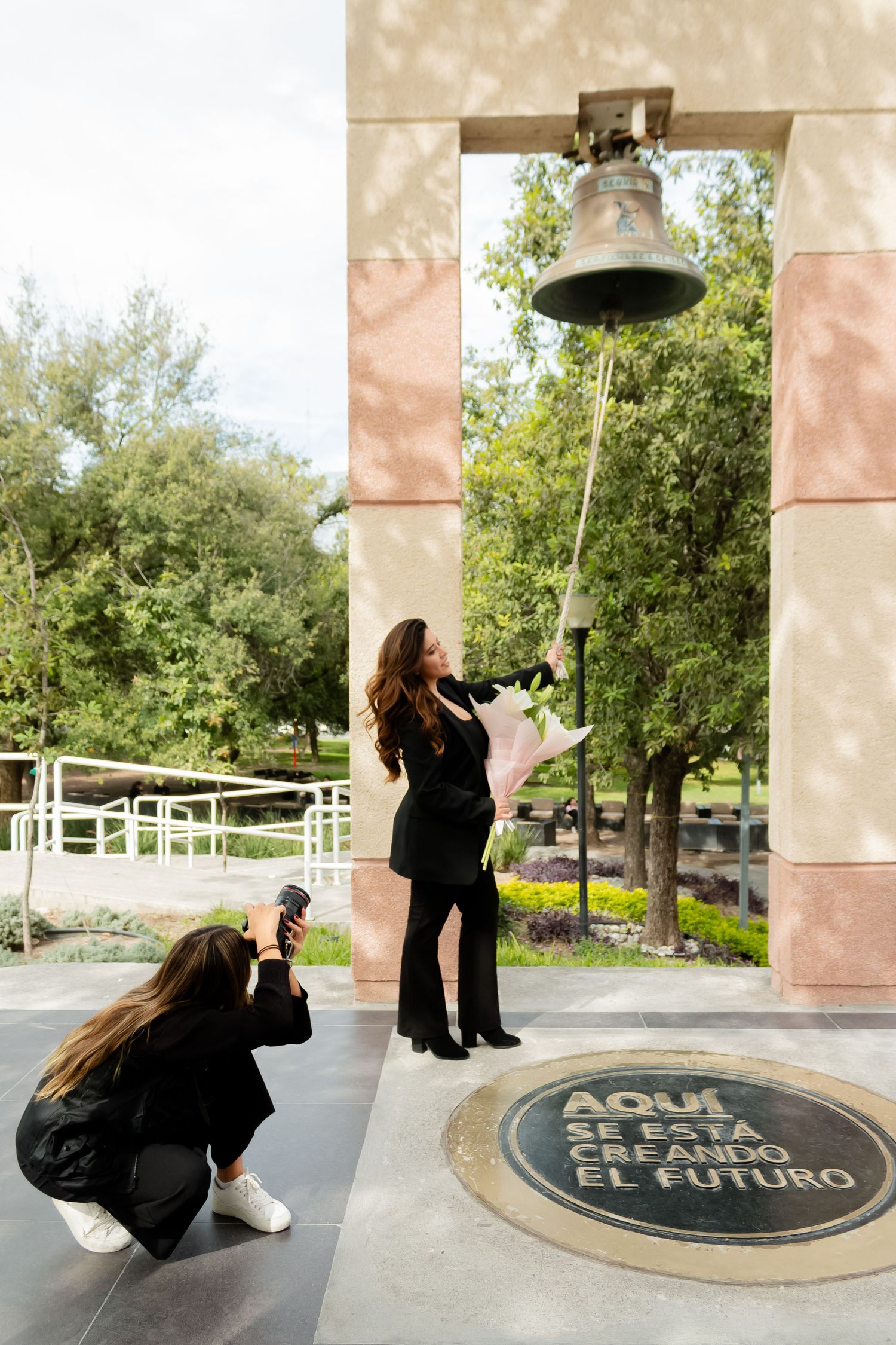 Graduate in black outfit rings a ceremonial bell while a friend photographs the milestone moment.