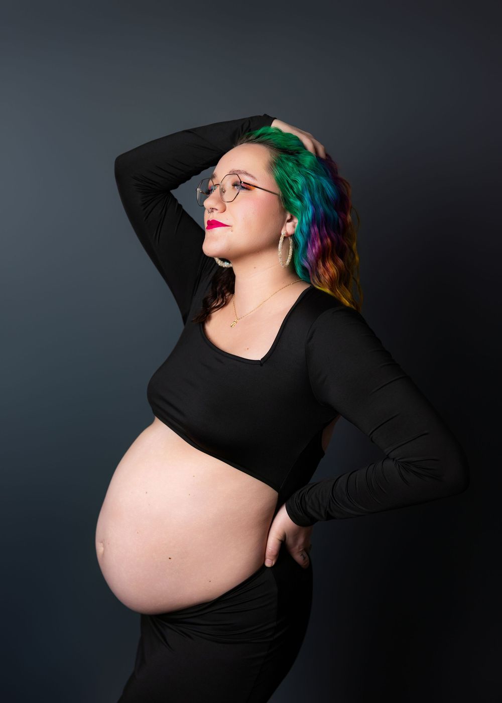 Maternity portrait featuring vibrant rainbow-colored hair against dark background wearing a black crop top.