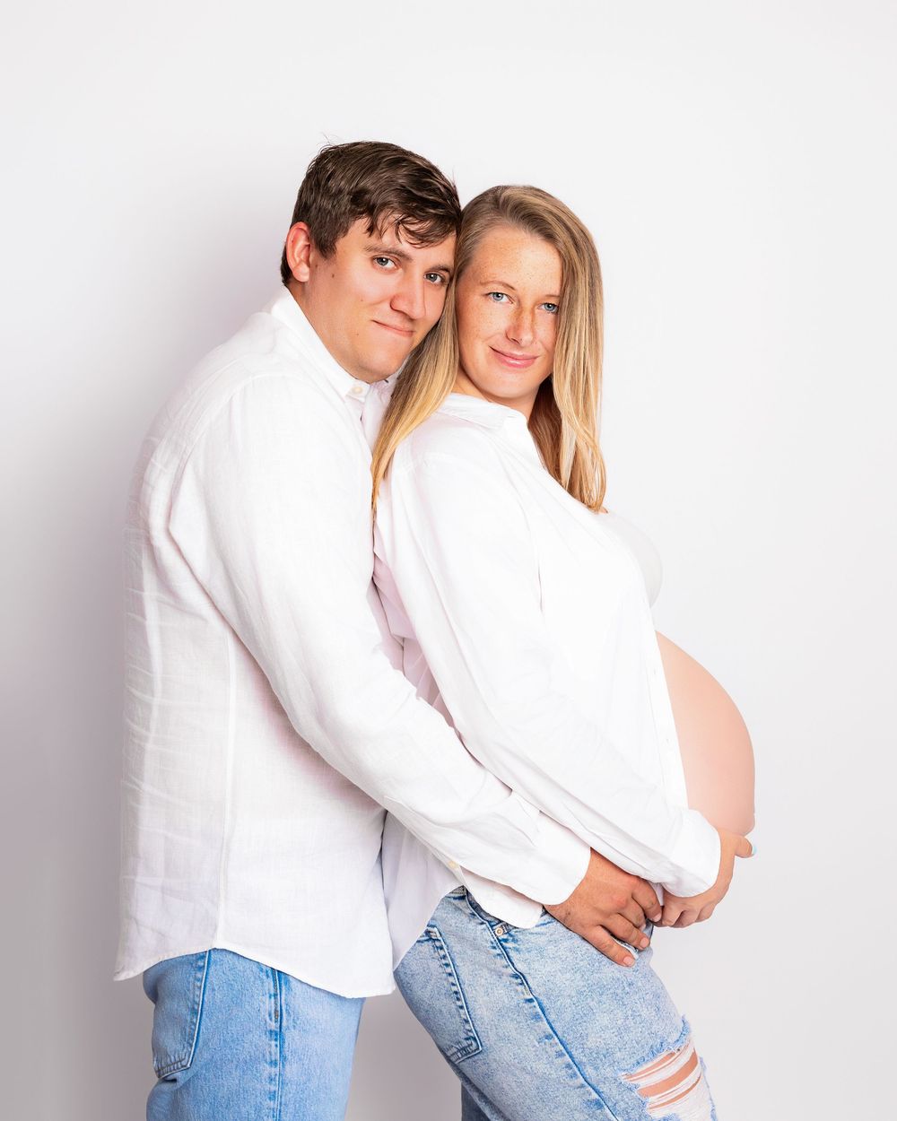 Couple in white shirts and jeans pose together for maternity photo against white background.