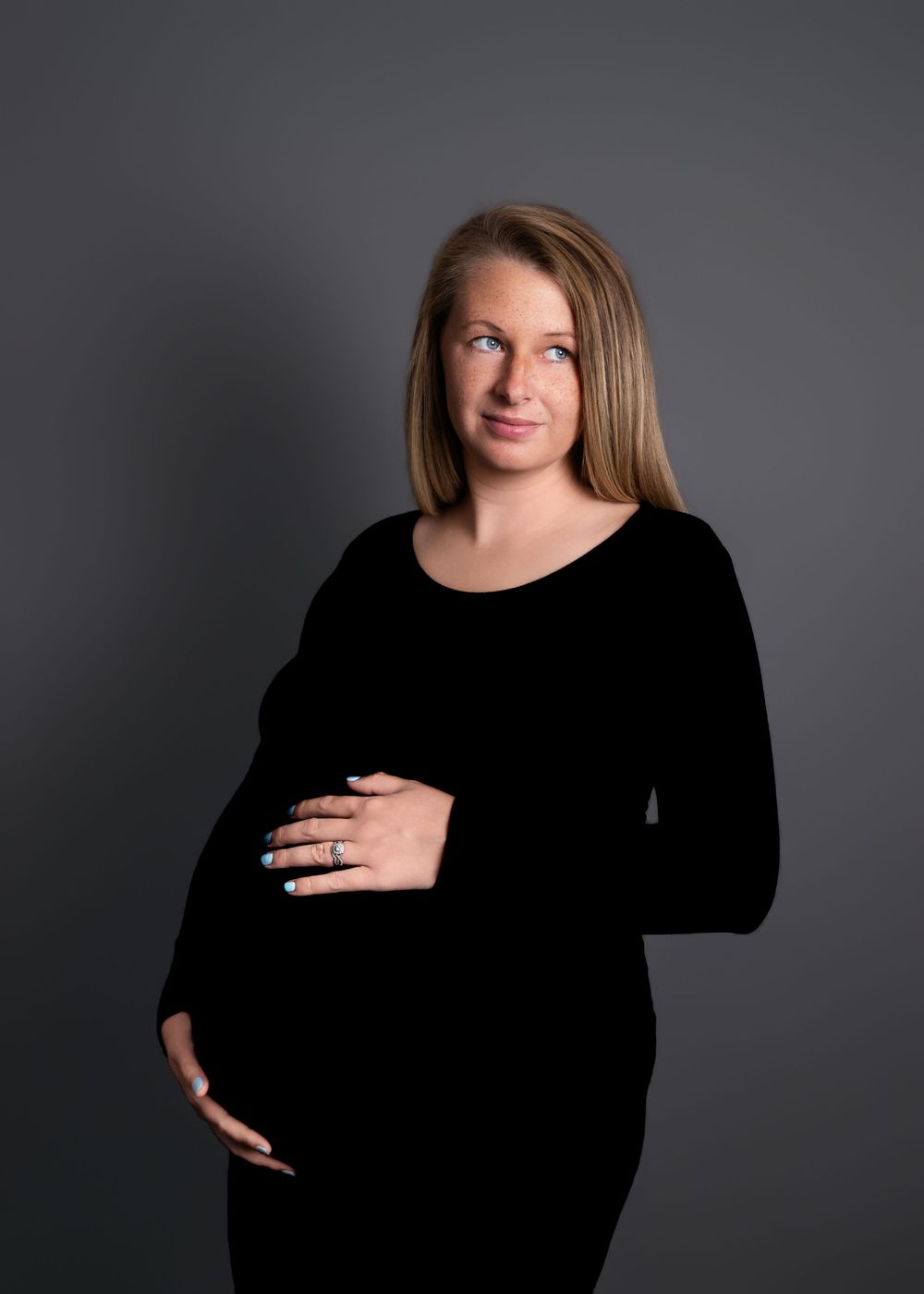 Studio maternity portrait of an expectant mother in a black dress against a dark gray background.