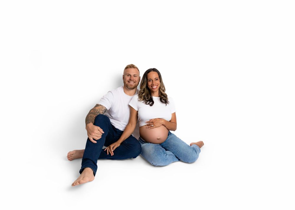 A couple sits on a white backdrop during a maternity photo shoot, with the expecting mother wearing jeans and cradling her belly.