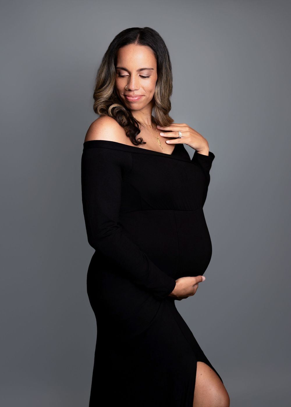 Elegant maternity photo of woman in black off-shoulder dress against grey backdrop, touching neckline with closed eyes.