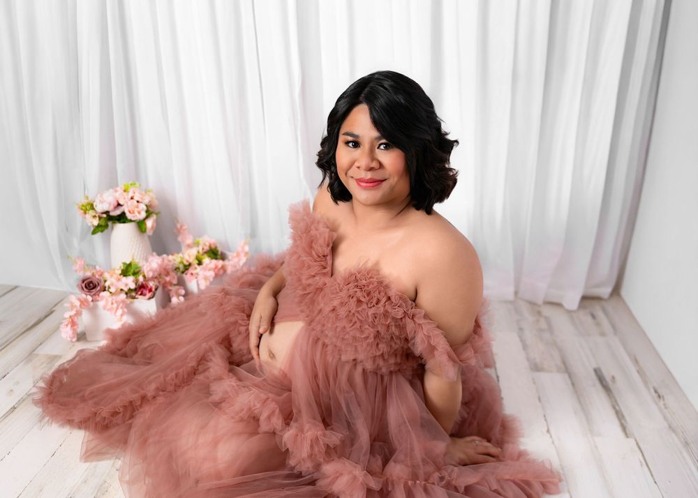 A woman in a dusty rose tulle gown poses elegantly on a white floor with floral arrangements in the background.