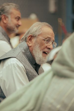 An elderly man in glasses and a grey vest smiles while engaged in conversation.