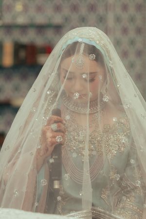 A bride wears an ornate embellished veil and traditional South Asian bridal attire.
