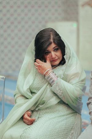 A bride in a light colored traditional outfit wipes away tears during an emotional moment.