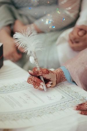 Signing ceremony detail with henna-decorated hands holding feather pen over marriage document.