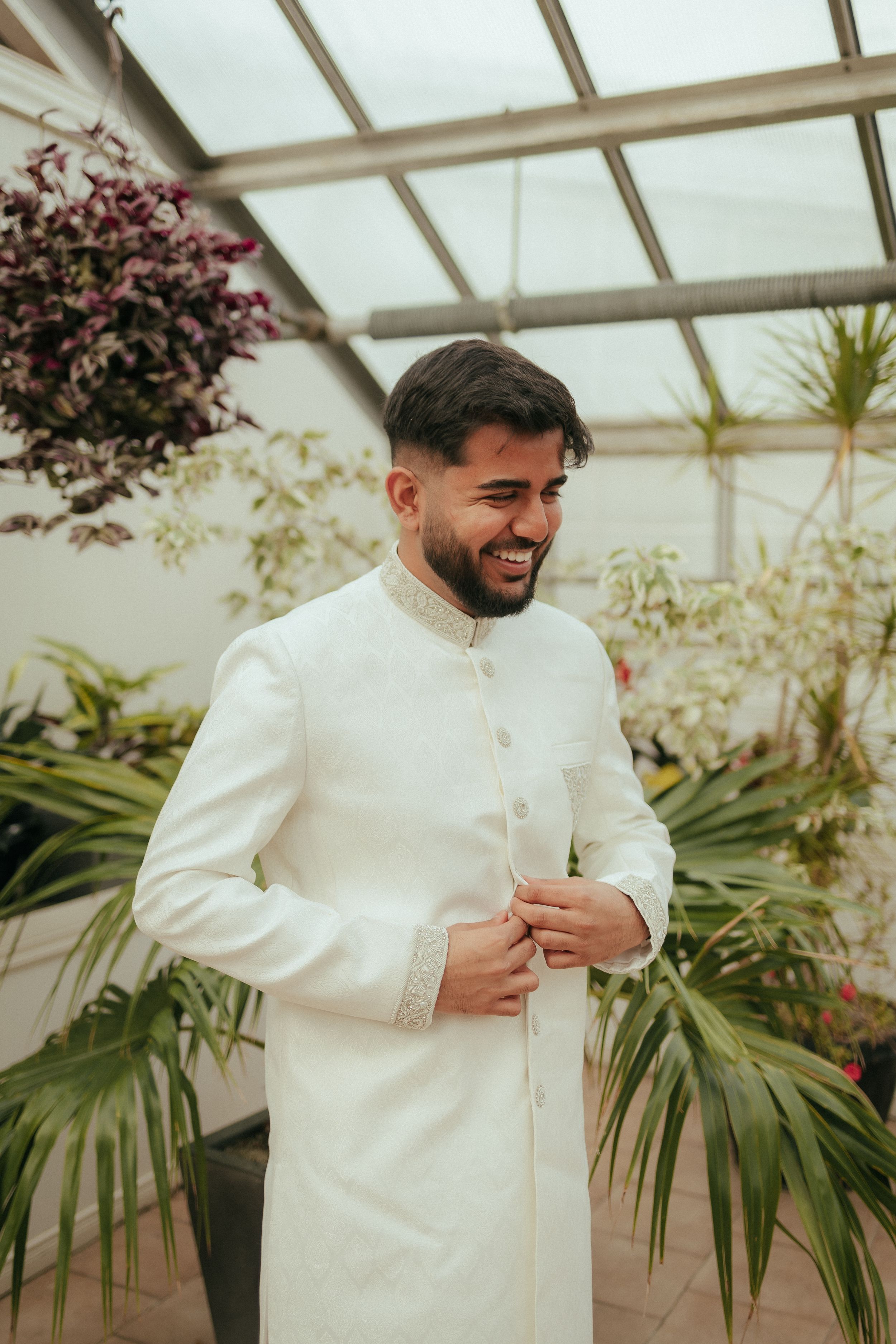 Groom wearing traditional white sherwani smiles while adjusting his collar in a sunlit greenhouse setting.
